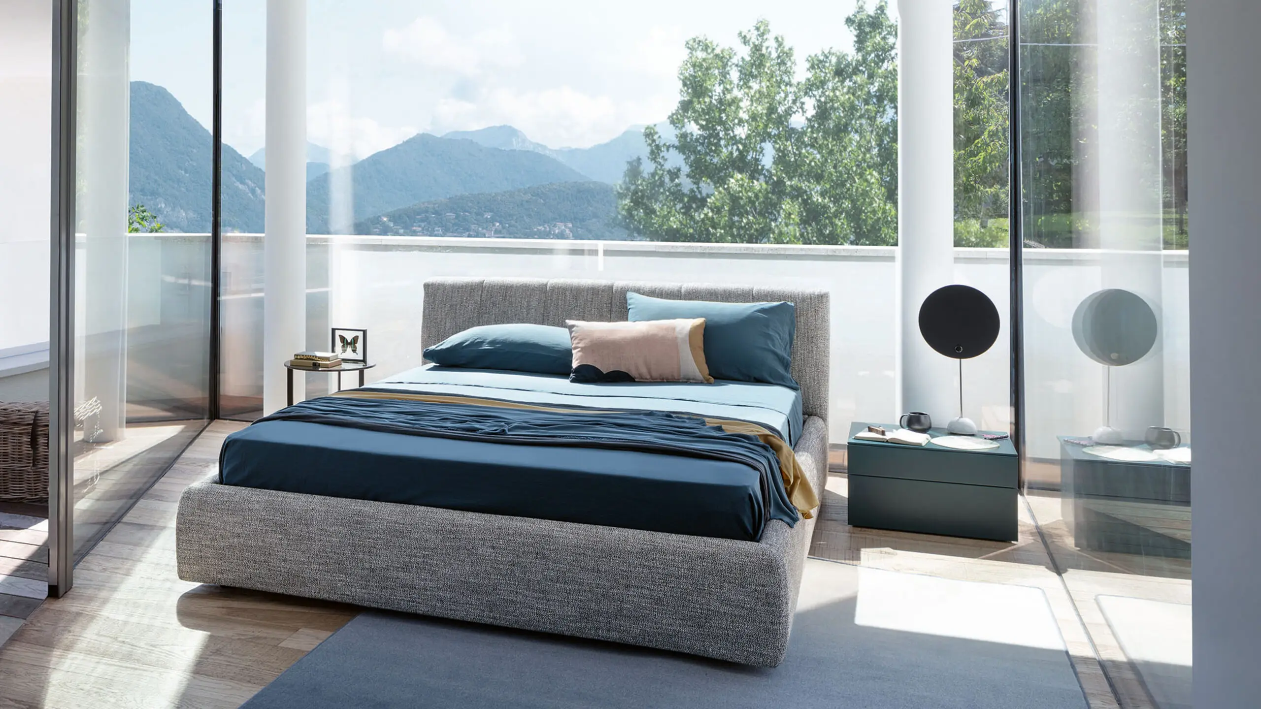 Camera con letto imbottito in tessuto grigio, lenzuola blu, comodino verde scuro e vetrate affacciate su monti e alberi